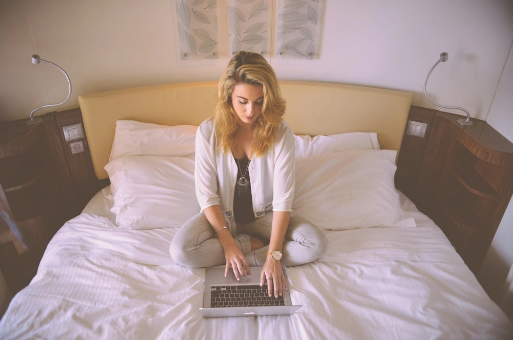 kobieta siedząca po turecku na hotelowym łóżku i patrząca na laptopa